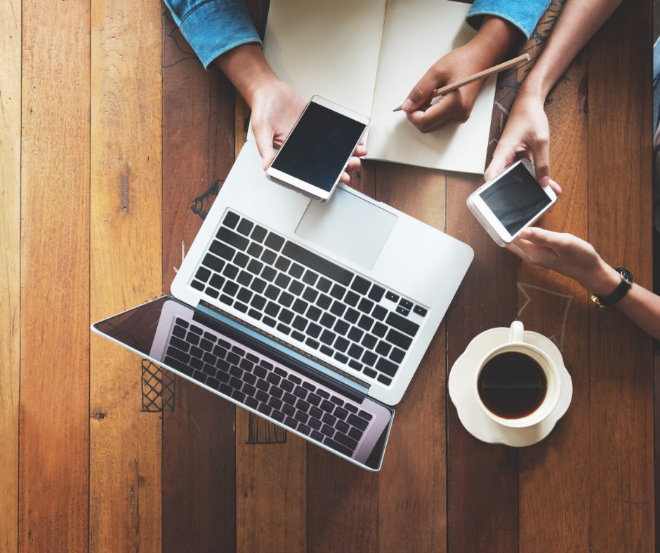 Two people's hands. Each person has a cell phone in their hand and there is one laptop sitting on a table