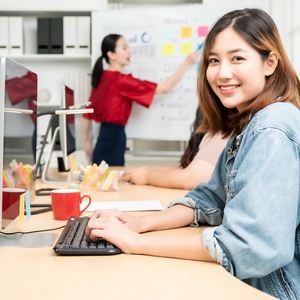 A smiling leader typing on her computer with a coworker working on a flow chart in the background